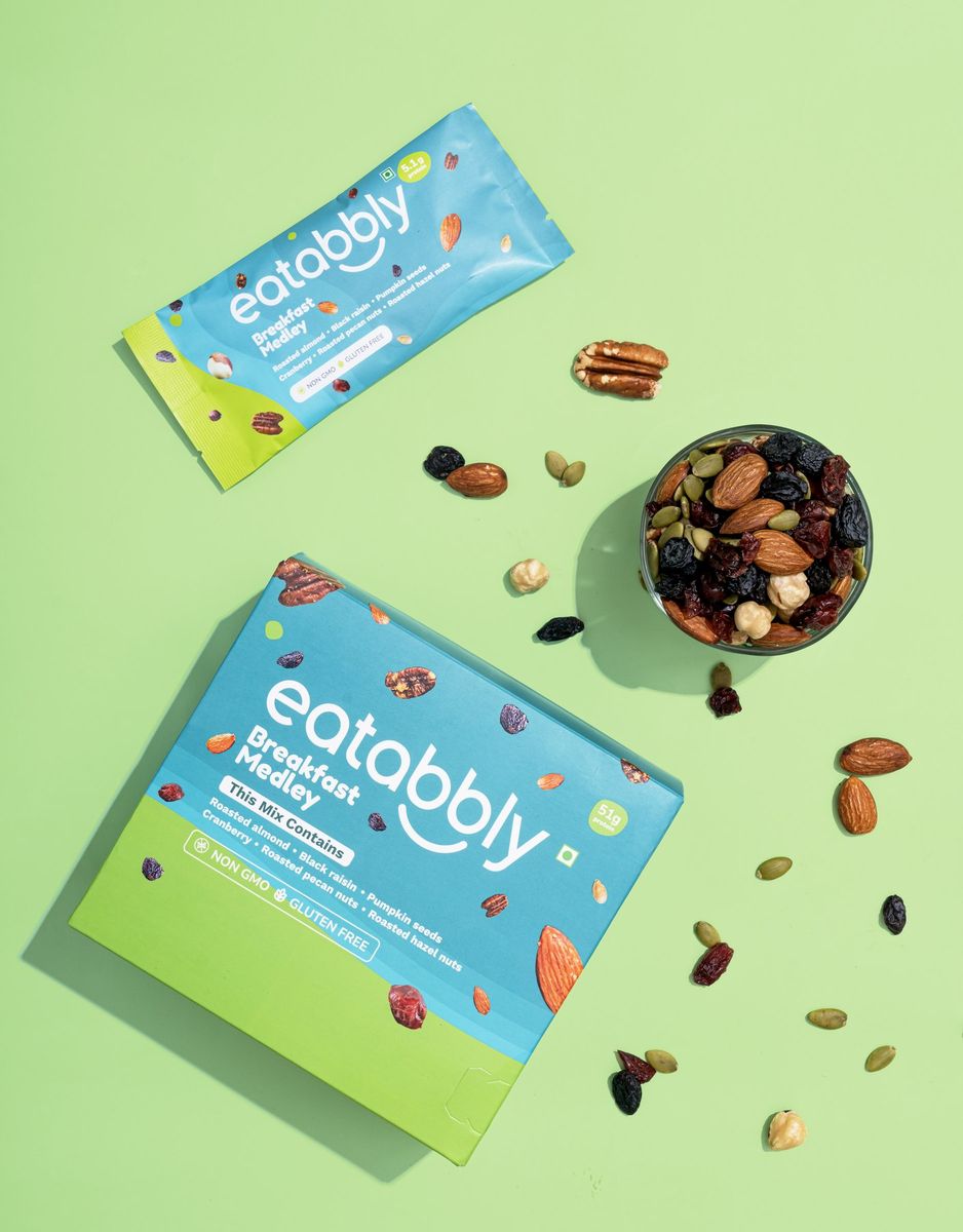 An Eatabbly Breakfast Medley pouch and box on a green background, with a bowl of the mixed nuts and loose almonds, pecans and raisins around them