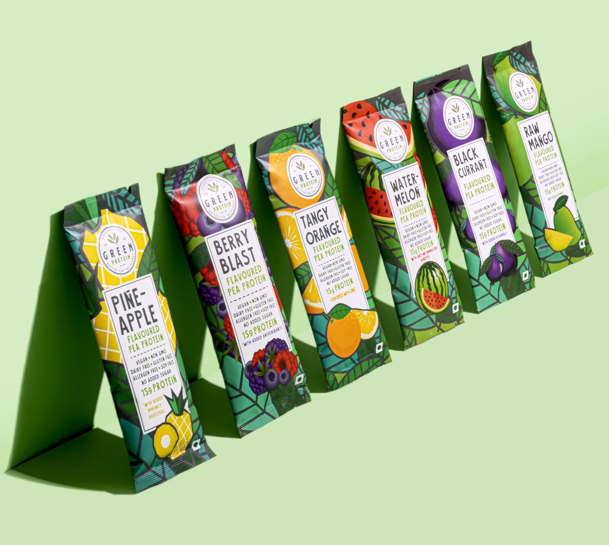 Six flavours of Green Protein pea protein sachets lined up diagonally on a pale green background, each flavour name readable on the front of the pack